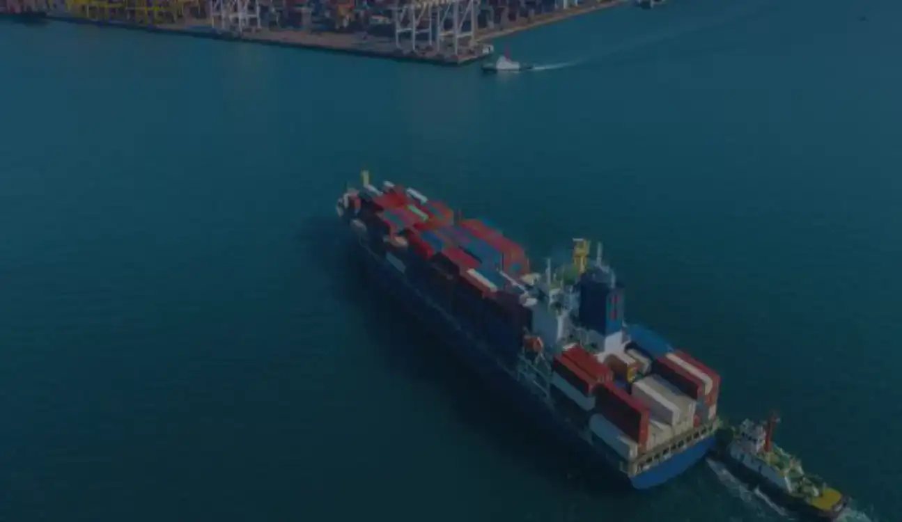 Container ship sailing in port waters with escort vessel, cranes and container yard in background